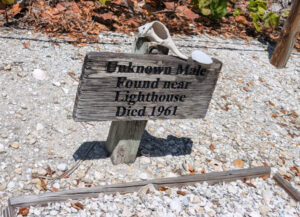 Wooden grave marker with seashells in Sanibel, Florida reading ‘Unknown Male Found Near Lighthouse Died 1961’ in a sandy setting